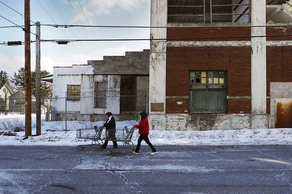 Women push shopping carts past the abandoned Surpas Leather Company that was built in 1920. Remnants of Gloversville’s leather tanning industry are spread throughout the city’s neighborhoods. Image by Larry C. Price. United States, 2016.