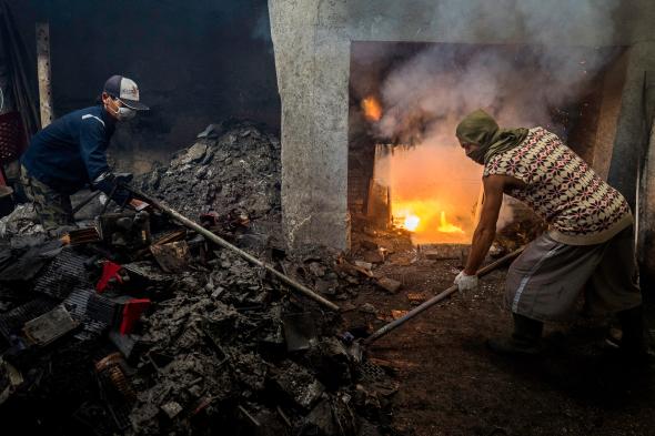 Two workers shovel lead battery cells into the fire at the small Garuda Jaya smelter in Kebasen as smoke pours into the room. Soot from the recycling operation drifts over nearby rice fields and villages, where residents complain that it burns their eyes, makes them dizzy, and gives them headaches. Image by Larry C. Price. Indonesia, 2016.